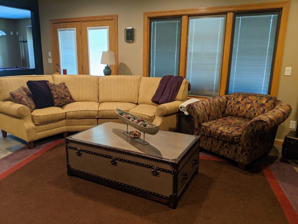 Before image of the space with gold colored angled sectional, patterned club chair, color block rug, and trunk style coffee table.
